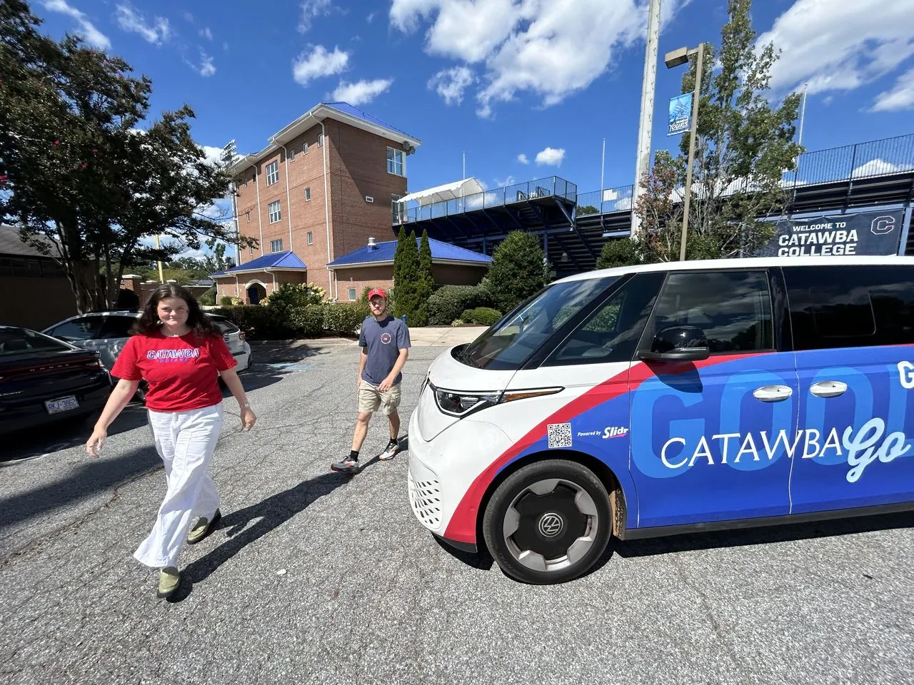 Two Catawba College students walking past a wrapped CatawbaGO electric shuttle parked on campus.